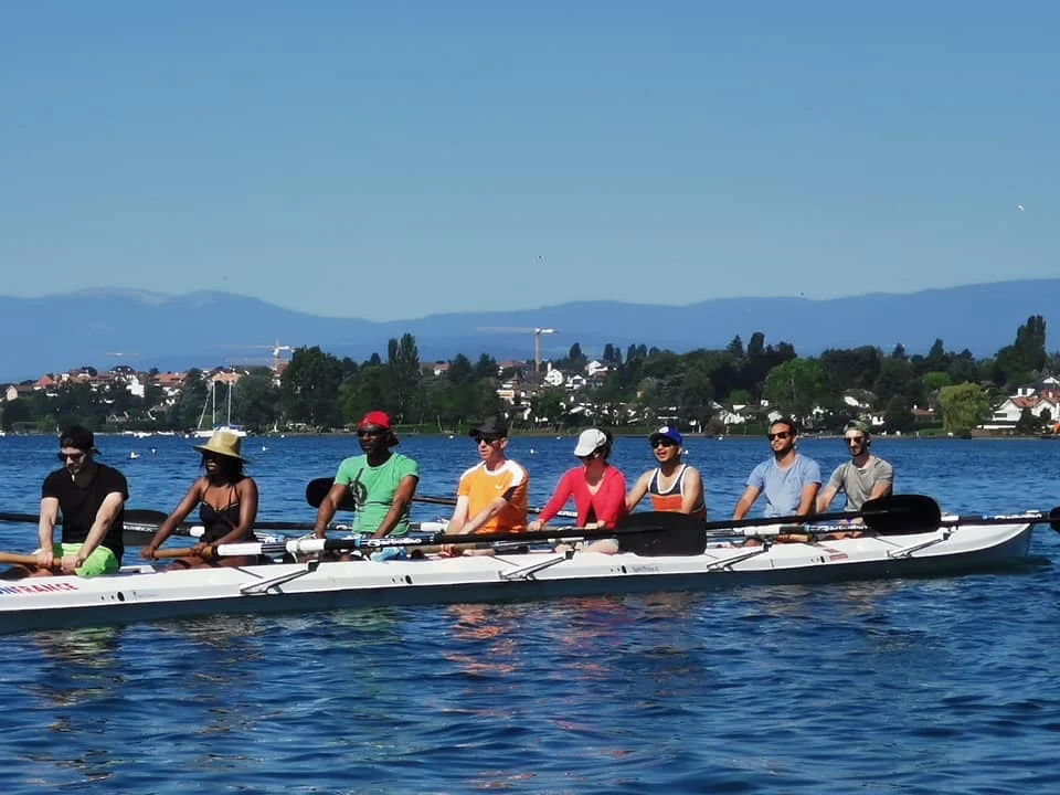 A crew in colourful kit rows in the sunshine on Lake Geneva